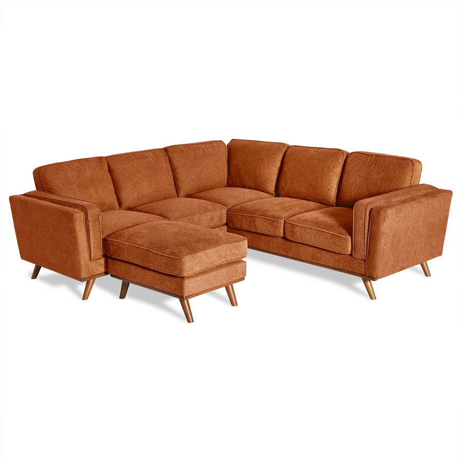 A spacious loft with brick walls, large windows, and concrete floors features an orange sectional sofa, a circular white rug, a wooden desk, a chair, a plant, and a set of shelves in the background.