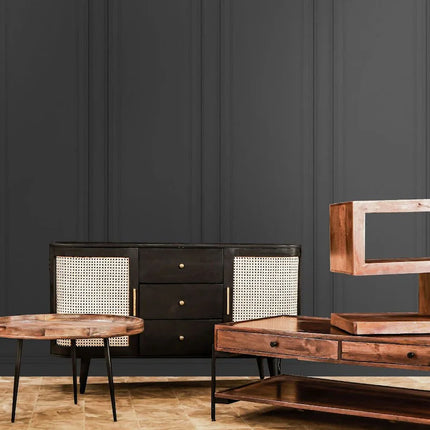 Minimalist furniture set in a room with dark panel walls. Includes a sideboard with cane detailing, a long coffee table with a geometric sculpture, and round side tables, all in wood and metal.