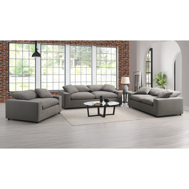 A modern living room showcases high-quality Peyton Classic Sofas in a U-shape around a round coffee table. A floor lamp enhances the natural light from large windows against a brick wall, while a potted plant adorns the wooden floor corner.