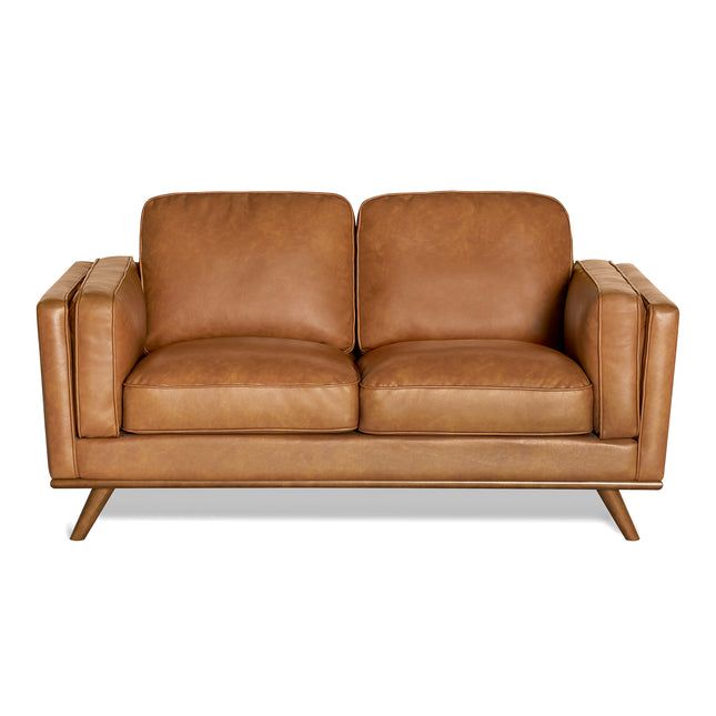 The Maddox Loveseat is a brown leather piece with plush cushions and a mid-century modern design, featuring two seat and back cushions, slightly angled wooden legs, and thick, boxy armrests that enhance its sleek look.