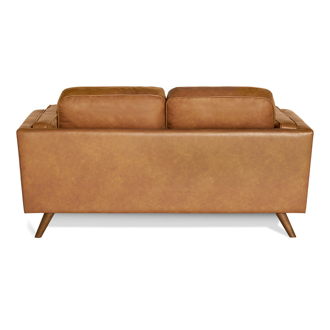 The Maddox Loveseat is a brown leather, mid-century modern piece showcased from the back with plush rectangular cushions and angled wooden legs, offering a sleek, minimalist design.
