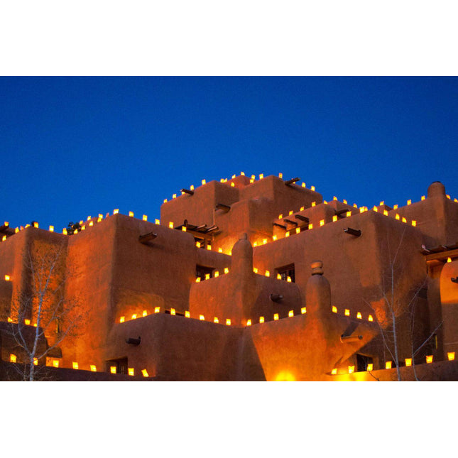 Illuminated by glowing lanterns, the Adobe Sante Fe building shimmers like museum-quality artwork under the deep blue sky. The warm light highlights its terracotta hue, creating a festive atmosphere with bare trees standing elegantly in the foreground.