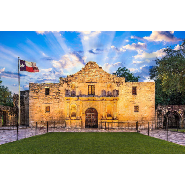 The image showcases the Alamo in San Antonio, Texas, at sunset. The illuminated building stands with a cloudy sky and sun rays above, a waving Texas flag on the left, and a green lawn in the front, creating museum-quality artwork.